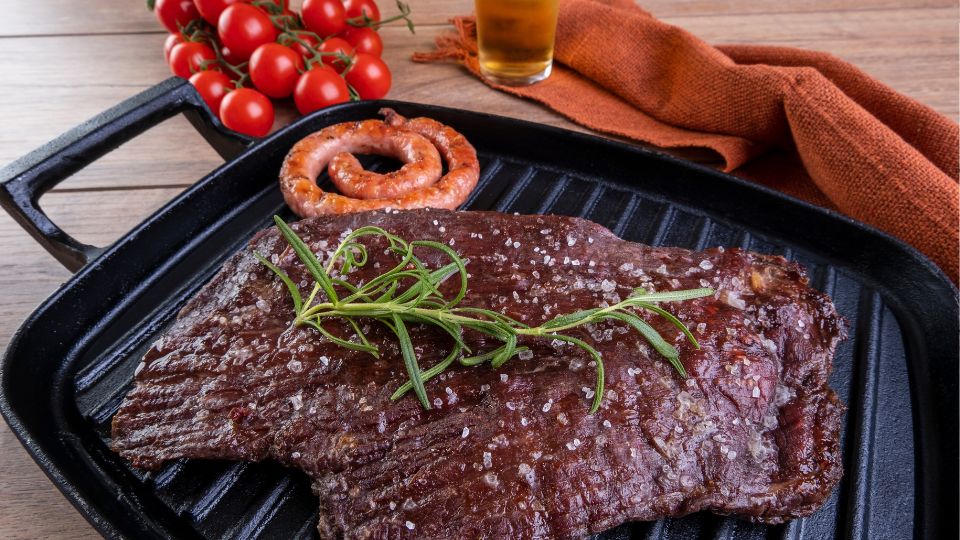Ein gegrilltes Steak mit Würstchen und frischen Tomaten auf einer Grillplatte.