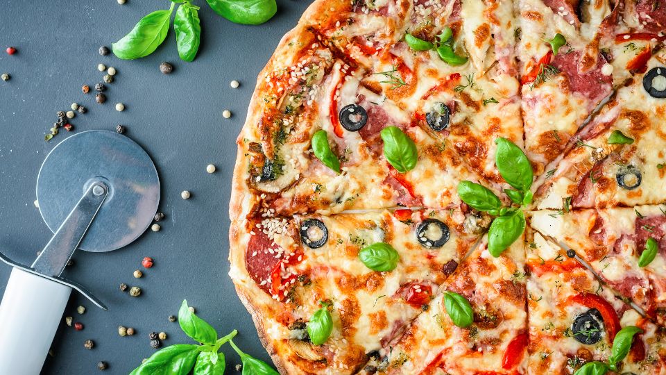 Eine frisch gebackene Pizza mit Basilikum, Oliven und Gewürzen auf einem Tisch.